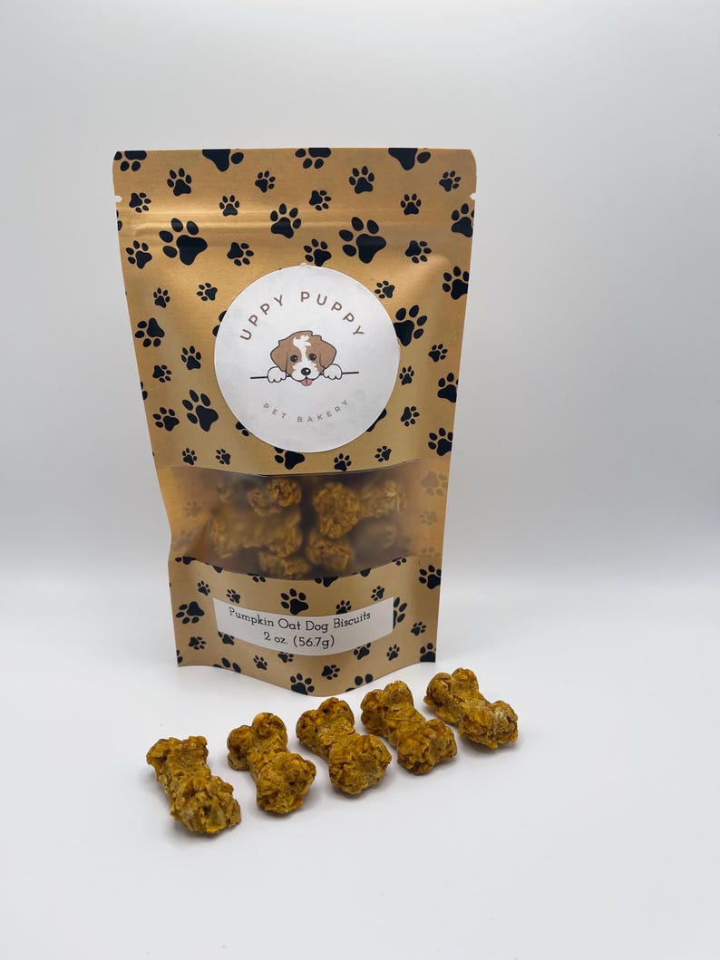 May include: A brown bag with a paw print pattern and the text "Uppy Puppy Pet Bakery" on the front. The bag contains pumpkin oat dog biscuits. There are six orange bone-shaped dog biscuits on a white surface.