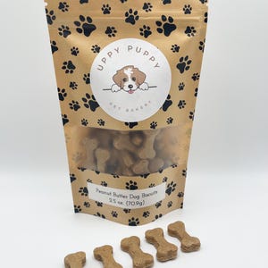May include: A brown paper bag with a paw print pattern and a logo that says "Uppy Puppy Pet Bakery". The bag contains peanut butter dog biscuits. There are five peanut butter dog biscuits in the shape of bones on a white surface.