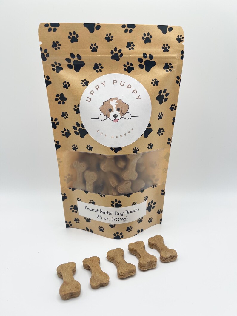 May include: A brown paper bag with a paw print pattern and a logo that says "Uppy Puppy Pet Bakery". The bag contains peanut butter dog biscuits. There are five peanut butter dog biscuits in the shape of bones on a white surface.