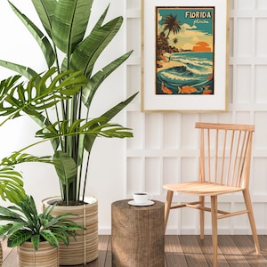 May include: A vintage-style poster with the text "FLORIDA" in a yellow banner, depicting a beach scene with palm trees, a sunset, and waves. The poster is framed in a light wood frame and hanging on a white wall.