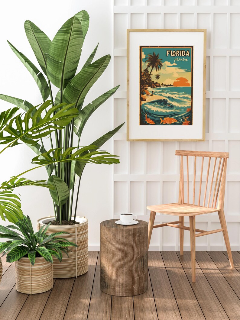 May include: A vintage-style poster with the text "FLORIDA" in a yellow banner, depicting a beach scene with palm trees, a sunset, and waves. The poster is framed in a light wood frame and hanging on a white wall.