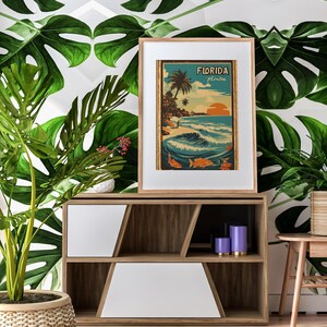 May include: A framed vintage travel poster with the text "Florida" and a depiction of a beach scene with palm trees, waves, and a surfer. The poster is displayed on a wooden shelf with a white front panel and purple candles.