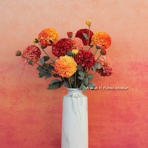 May include: A bouquet of orange, red and yellow dahlia flowers in a white marble vase against a gradient orange and red background. A and H Floral Studio.