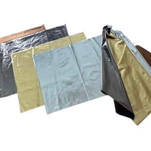 May include: An assortment of metallic fabric sheets in various colors, including bronze, charcoal, gold, silver, and pale yellow. The sheets are arranged in a layered, overlapping fashion, showcasing their reflective surfaces.