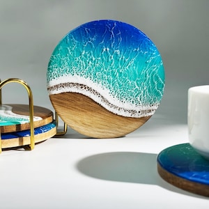 May include: Round wooden coasters with a resin ocean design. The coasters feature a gradient of blue and turquoise, with a white wave design and a brown wooden base. A set of coasters is in a gold metal holder.