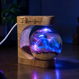 May include: A C-shaped wooden lamp base with the text I Love You engraved on the top, featuring a levitating clear glass sphere containing swirling blue and purple plasma light effects, powered by a white cord extending from the side of the base.