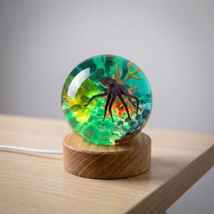 Puede incluir: Una lámpara esférica de resina con una figura de pulpo de color morado oscuro suspendida en un entorno acuático verde translúcido, con detalles de coral y rocas, montada sobre una base circular de madera clara con un cable blanco.