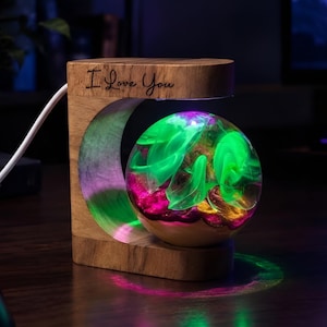 Könnte beinhalten: Ein C-förmiger Lampensockel aus Holz mit der Gravur I Love You und einer schwebenden, kugelförmigen Harzkugel. Die Kugel enthält wirbelnde grüne und magentafarbene Muster, die von internen LEDs beleuchtet werden und farbige Reflexionen werfen.