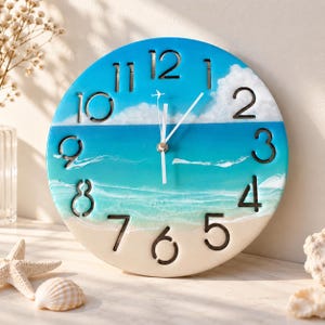 May include: A round wall clock featuring a beach and ocean scene with a blue sky, white clouds, turquoise water, and sandy shore. The clock face has large black numerals from 1 to 12, white clock hands, and a small white airplane icon at the 12.
