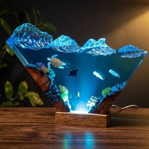 May include: A triangular resin lamp featuring a deep blue ocean scene with a sea turtle, a manta ray, two jellyfish, and a scuba diver. The top edge mimics crashing waves, while the base is made of natural wood with an integrated light source.