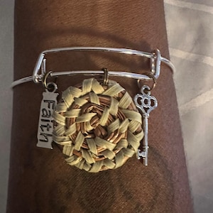 May include: Silver adjustable bracelet featuring a woven circular charm in beige and brown tones. Includes a key charm and a rectangular charm with the word "Faith". A fashionable accessory.