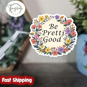 May include: A circular sticker with a floral border in shades of pink, yellow, orange, and purple. The sticker has the words "Be Pretti Good" in a black, elegant font. The sticker is being held by a pair of silver tweezers.