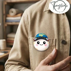 May include: A round, smiling cartoon character pin with a blue and green Earth-themed hat and rocket ship accents. The pin is attached to a tan jacket. The pin has the text "Thaylinh Design" in a circle.