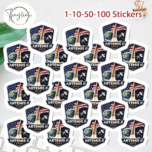 以下が含まれることがあります： 「ARTEMIS II 2026」の文字が入ったロケット発射のデザインのステッカーのコレクション。ステッカーは六角形で、青、白、赤の配色で、地球とアメリカ国旗が描かれています。