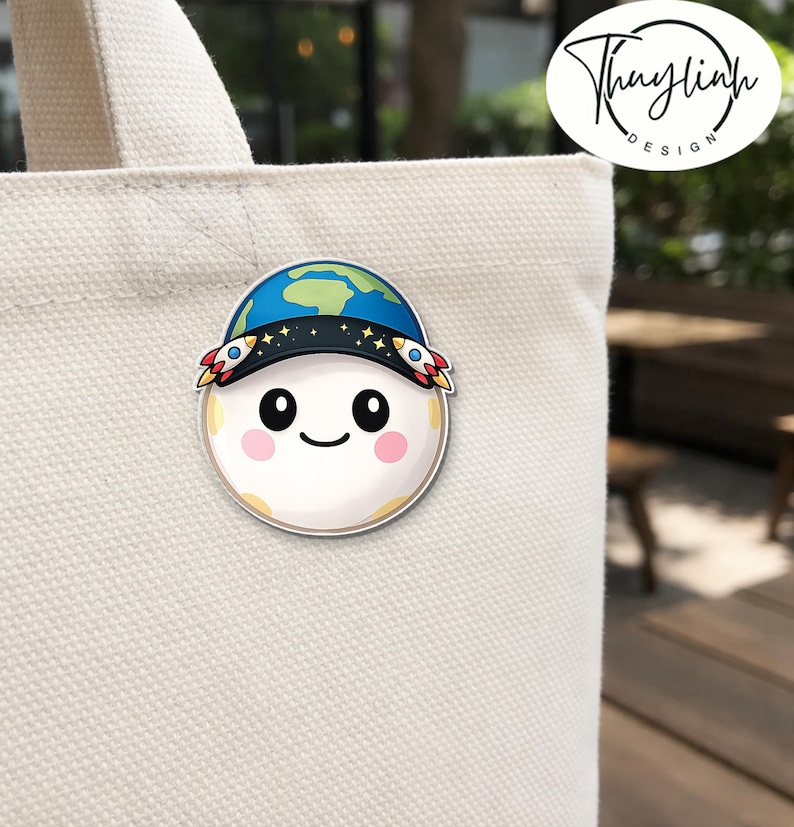 May include: A round sticker featuring a smiling moon wearing a blue and green Earth hat with rocket ship accents. The sticker is attached to a beige tote bag. The sticker has a white border and the moon has rosy cheeks.