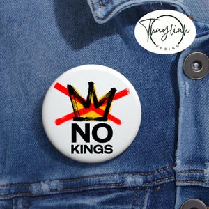 May include: A white button with the words "NO KINGS" in black, with a black and gold crown crossed out in red. The button is on a blue denim background. A smaller white button with a logo is also visible.