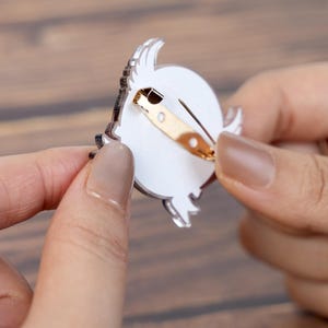 May include: A clear acrylic pin with a gold-colored clasp. The pin features a white circular design with a black outline and a decorative element. The pin is held between two hands.