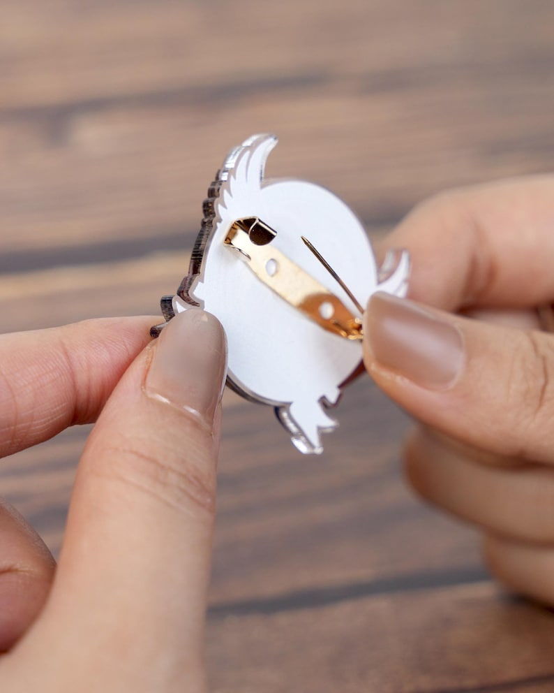 May include: A clear acrylic pin with a gold-colored clasp. The pin features a white circular design with a black outline and a decorative element. The pin is held between two hands.
