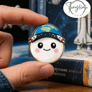 May include: A round enamel pin featuring a smiling moon character wearing a blue and green Earth hat with rocket ship accents. The pin has pink cheeks, black eyes, and a yellow border. The pin is held between fingers.