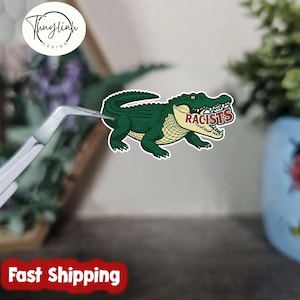 May include: A green alligator sticker with the word "RACISTS" in its mouth. The sticker is held by tweezers. The sticker has a white border and a yellow underbelly. The sticker is a cartoon illustration.
