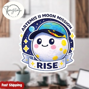 Puede incluir: Una pegatina con una luna de dibujos animados que lleva una gorra con un diseño de planeta. La pegatina tiene el texto "ARTEMIS II MOON MISSION" y "RISE" en una pancarta, con estrellas y un fondo azul.