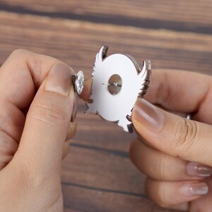 May include: A silver-colored owl-shaped pin with a white center and brown outline. The pin has a metal clasp on the back for secure attachment. The pin is being held up by a person's hands.