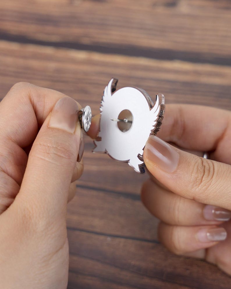 May include: A silver-colored owl-shaped pin with a white center and brown outline. The pin has a metal clasp on the back for secure attachment. The pin is being held up by a person's hands.