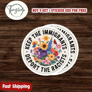 May include: Two round stickers with the text "KEEP THE IMMIGRANTS, DEPORT THE RACISTS" encircling a floral arrangement. The flowers include sunflowers, roses, and butterflies. The stickers are on a dark wood background.