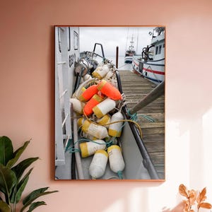 May include: A close-up of a fishing boat docked at a pier. The boat is filled with orange and white buoys, and there are two other boats in the background. The pier is made of wood and is weathered.