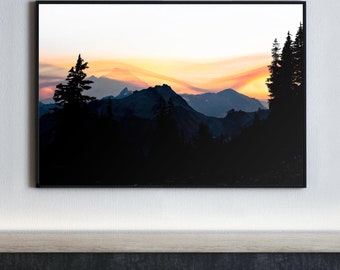 May include: A black framed photograph of a mountain range silhouetted against a sunset sky. The sky is a gradient of orange, yellow, and pink. The mountains are dark and rugged, with a few trees visible in the foreground.