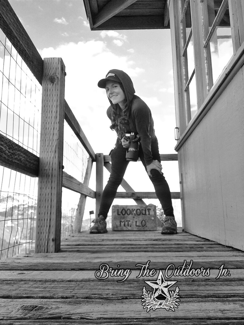May include: A person wearing a black hooded sweatshirt and a black baseball cap stands on a wooden platform with a sign that reads "Lookout Mt. Lo."