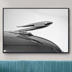 Può includere: Una fotografia in bianco e nero di un ornamento per il cofano di un'auto d'epoca. L'ornamento è un uccello cromato in volo, con un lungo becco appuntito e le ali spiegate. L'ornamento è montato su un cofano di un'auto nera.