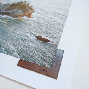 May include: A close-up of a white mat with a brown ruler on the bottom right corner. The ruler is labeled "X-ACTO" and "PRECISION EDGE". The mat has a photograph of a rocky island in the ocean on it.
