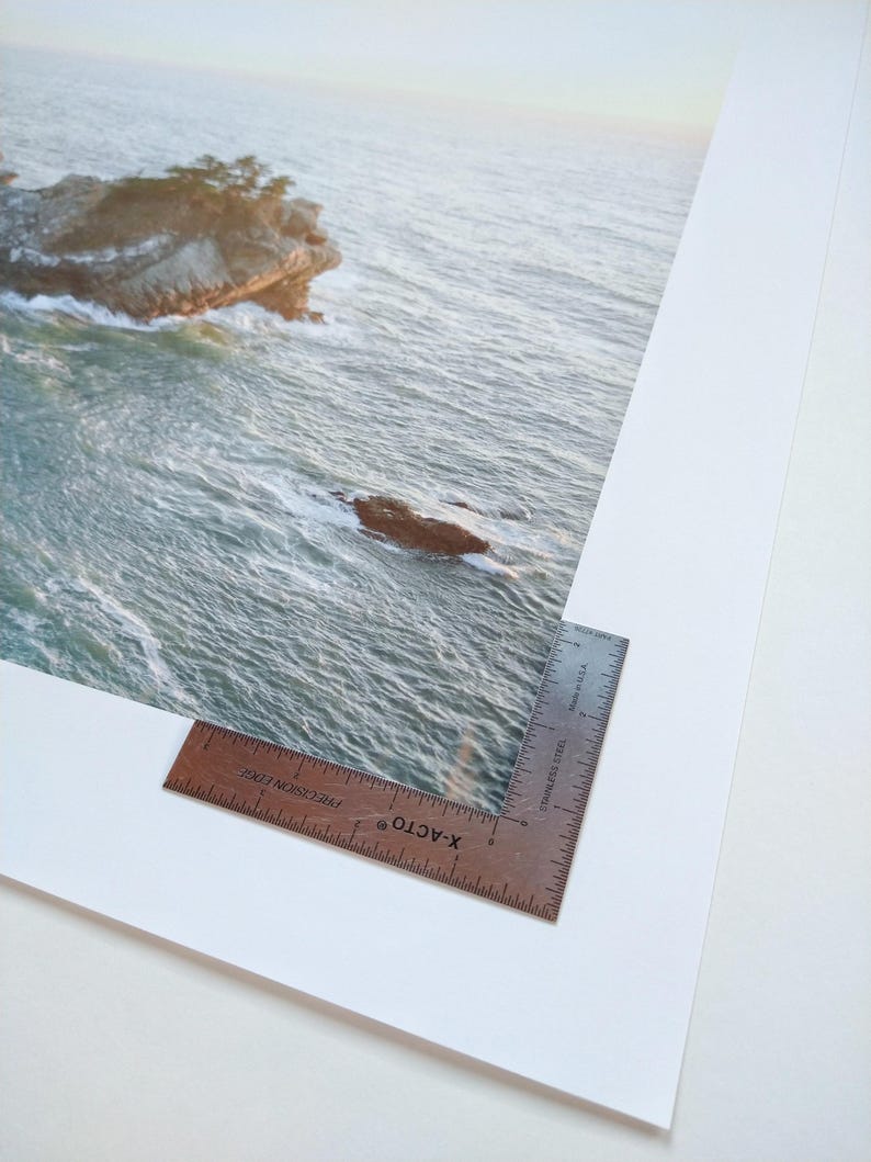 May include: A close-up of a white mat with a brown ruler on the bottom right corner. The ruler is labeled "X-ACTO" and "PRECISION EDGE". The mat has a photograph of a rocky island in the ocean on it.
