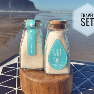 May include: Two small glass jars with cork lids filled with white salt. The jars have blue and white labels that say "Wild Coast" and "Wet Salt Handmade on the Oregon Coast 4 ounces". The jars are sitting on a wooden platform with a blue and white tiled background. The text "TRAVEL SET" is in a white hexagon on a blue background.