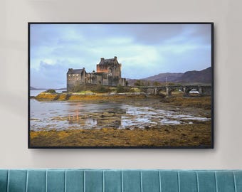 May include: A framed print of Eilean Donan Castle in Scotland. The castle is a stone structure with multiple towers, set on a small island. A stone bridge connects the castle to the mainland. The sky is overcast, and the water reflects the castle.