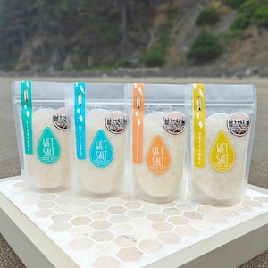 Puede incluir: Cuatro bolsas transparentes de sal húmeda, cada una con una etiqueta de diferente color: turquesa, azul, naranja y amarillo. Cada etiqueta tiene las palabras "Wet Salt" y un nombre único. Las bolsas se muestran sobre una superficie de baldosas decorativas.