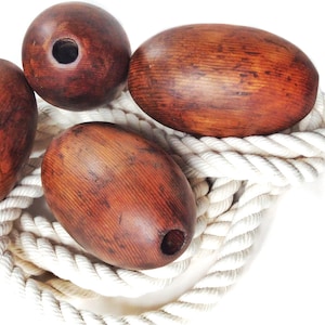 Puede incluir: Cuatro boyas de madera en formas ovaladas y redondas, cada una con un agujero. Las boyas son de color marrón cálido con vetas de madera visibles. Descansan sobre una gruesa cuerda blanca trenzada, creando un tema náutico.