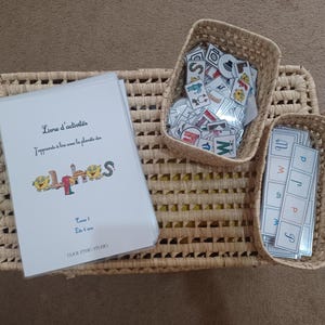 May include: A wicker basket tray with two smaller wicker baskets. The larger basket contains a book titled "Livre d'activités J'apprends à lire avec la planète des alphas" and the smaller baskets contain letter tiles.