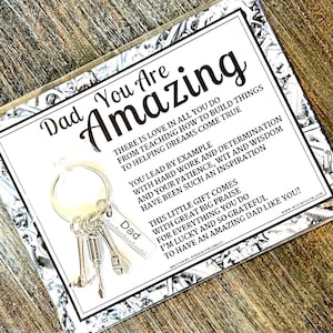 May include: A silver keychain with miniature tools and a tag that says "Dad" is displayed on a card. The card reads "Dad You Are Amazing" with a heartfelt message. The background is a wooden surface.