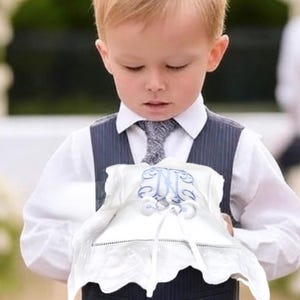 May include: A young boy in a navy blue pinstripe vest and pants, white shirt, and gray tie holds a white, scalloped pillow with blue embroidered initials. The pillow has a ribbon and is likely for a wedding.