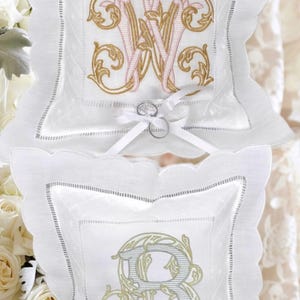 May include: Two white ring bearer pillows with scalloped edges. One pillow features a gold and pink embroidered monogram "W", the other has a gold and green embroidered "B". Each pillow has a white ribbon and holds two rings.