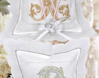 May include: Two white ring bearer pillows with scalloped edges. One pillow features a gold and pink embroidered monogram "W", the other has a gold and green embroidered "B". Each pillow has a white ribbon and holds two rings.