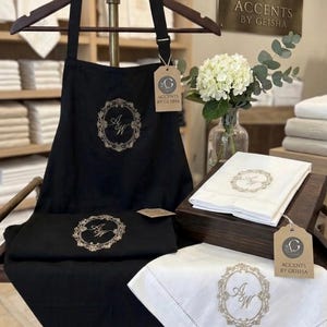 May include: Black apron and matching linens with embroidered floral wreath and initials. White linens with embroidered details are also displayed. The brand name "Accents by Geisha" is visible on a sign and tags.