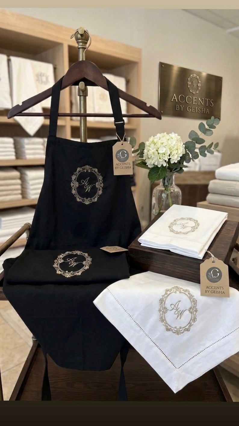 May include: Black apron and matching linens with embroidered floral wreath and initials. White linens with embroidered details are also displayed. The brand name "Accents by Geisha" is visible on a sign and tags.