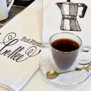 May include: White kitchen towel with the text "But First... Coffee" in black cursive lettering. A black and white illustration of a coffee pot is on a second white kitchen towel. A glass of coffee sits on a white doily with a gold spoon.