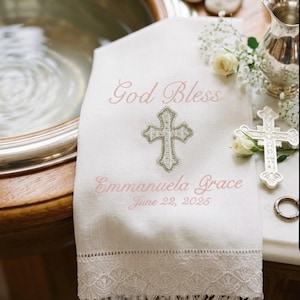 May include: White baptismal towel with embroidered text "God Bless", a cross, and the name "Emmanuela Grace" with the date "June 22, 2025". The towel has a delicate lace trim and is near a baptismal font.