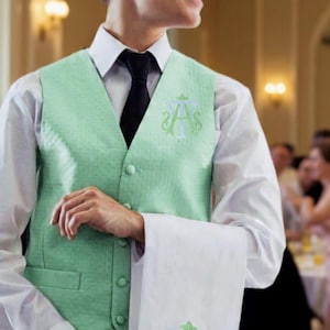 May include: A person wearing a light green vest with a white shirt and black tie. The vest and white towel feature an embroidered monogram. The setting appears to be a formal event.