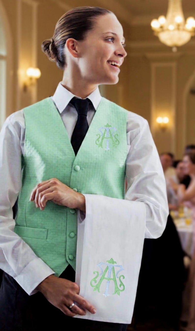 May include: A person wearing a light green vest with a white shirt and black tie. The vest and white towel feature an embroidered monogram. The setting appears to be a formal event.