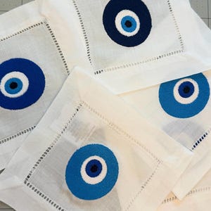 May include: Set of four white linen napkins with blue and white embroidered evil eye designs.